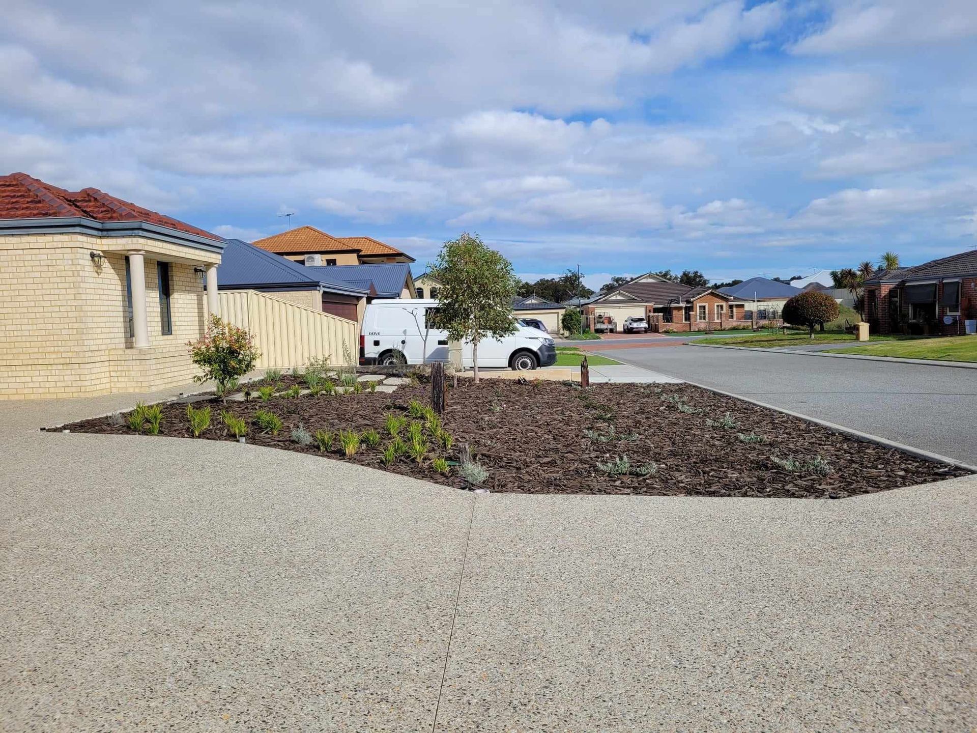 Residential street with a gravel driveway, landscaped bed, and houses under a cloudy sky.