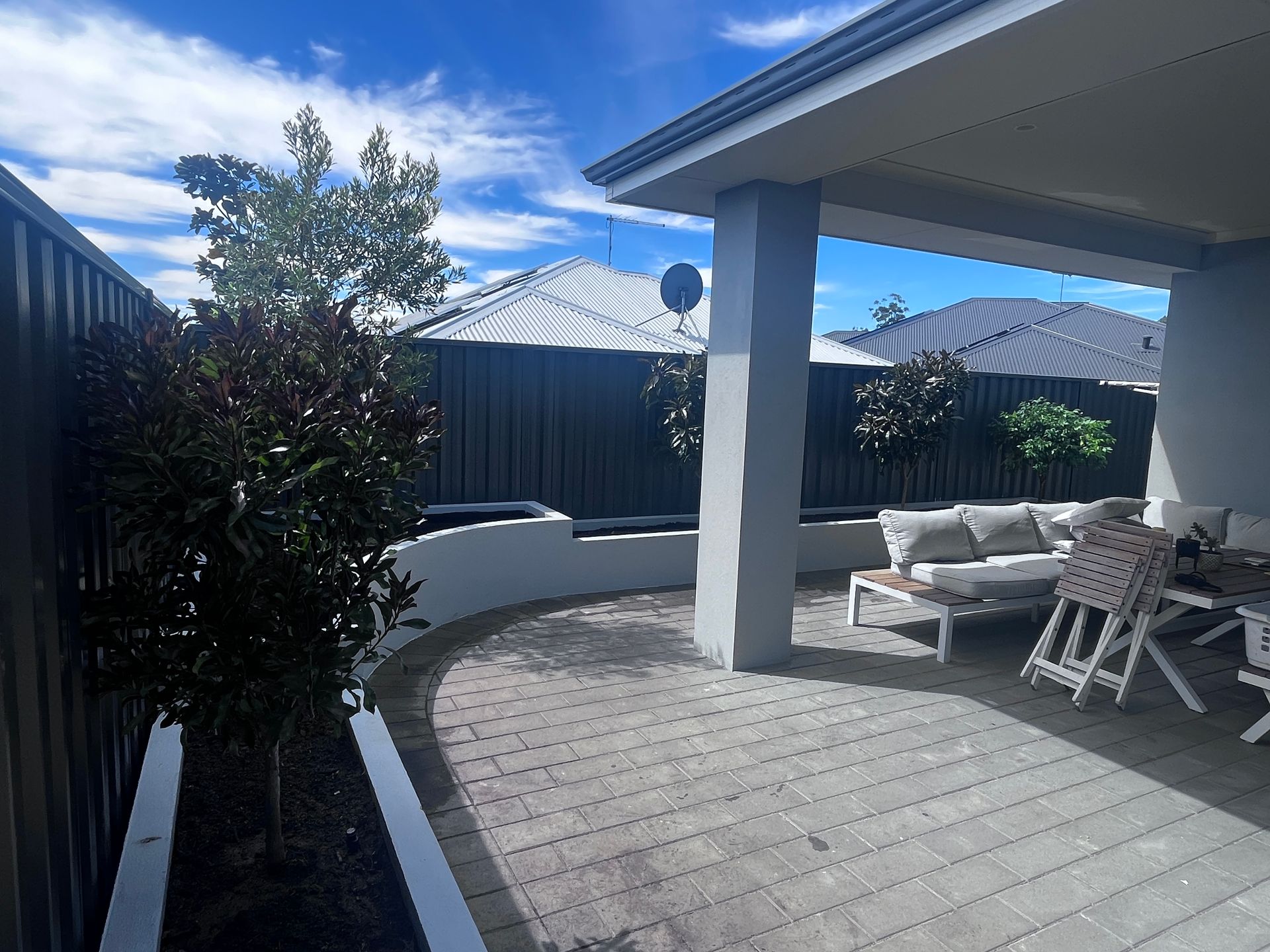 Backyard patio with paving, seating, and trees under a roof, bordered by a fence and a low wall.
