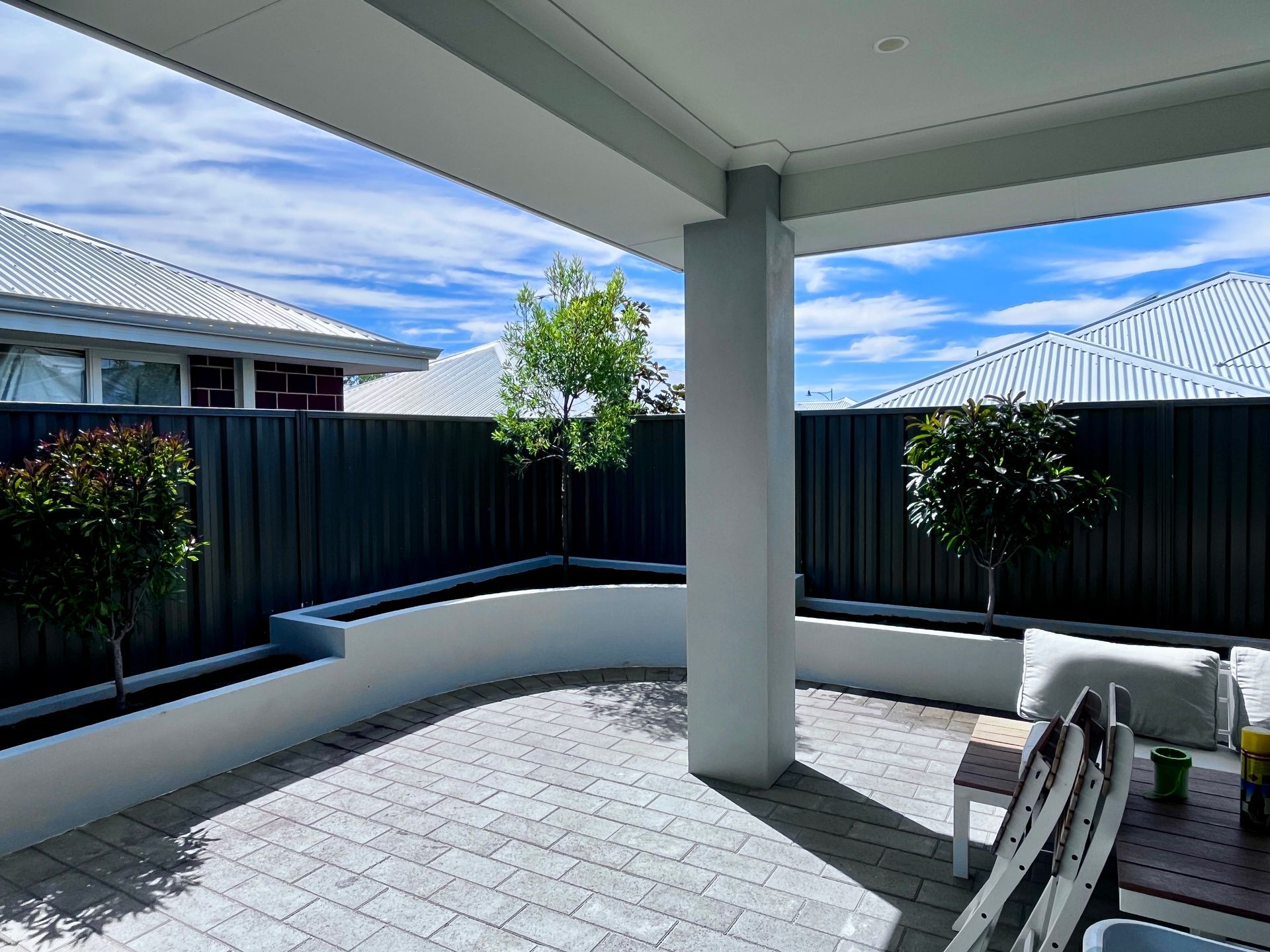 Patio with paved floor, white pillar, and low planters with small trees against a dark gray fence.