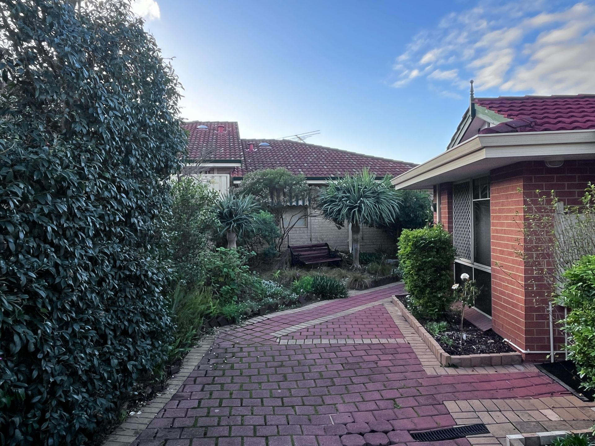 Brick pathway leading through a garden with a house on the right and other buildings in the background.