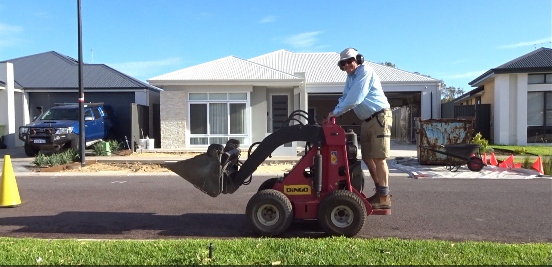A person operates a small red skid steer loader on a street in front of houses on a sunny day.