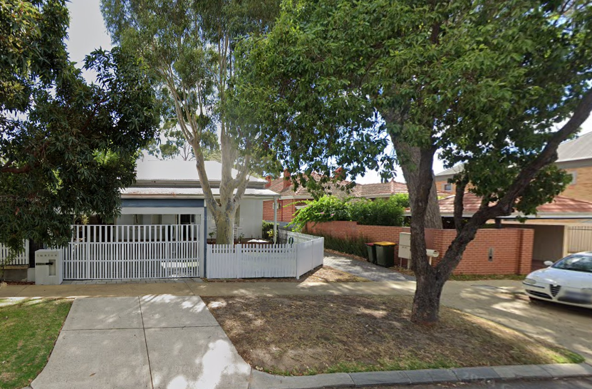 A single-story house with a white picket fence and trees on a sunny street.
