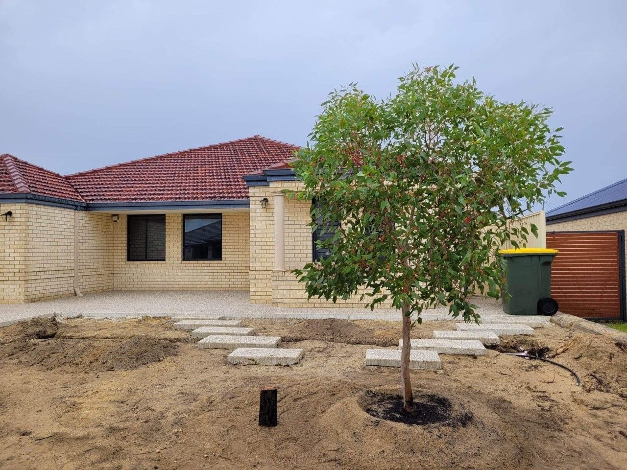 House exterior with a small tree in the foreground. Brickwork, patio, and a trash can are also visible.