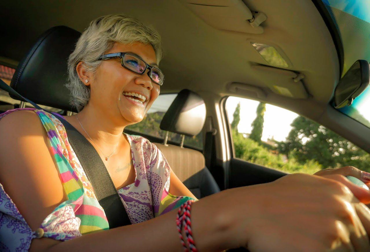 Woman with glasses smiles while driving a car; sunlight streams in.