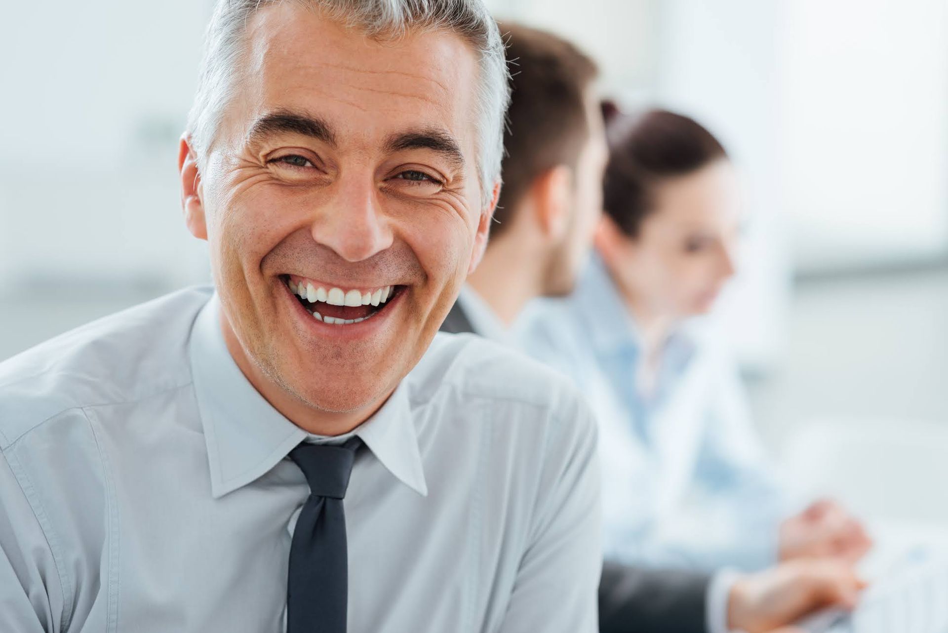 Smiling man in a light blue shirt and tie at an office desk, colleagues blurred in the background.