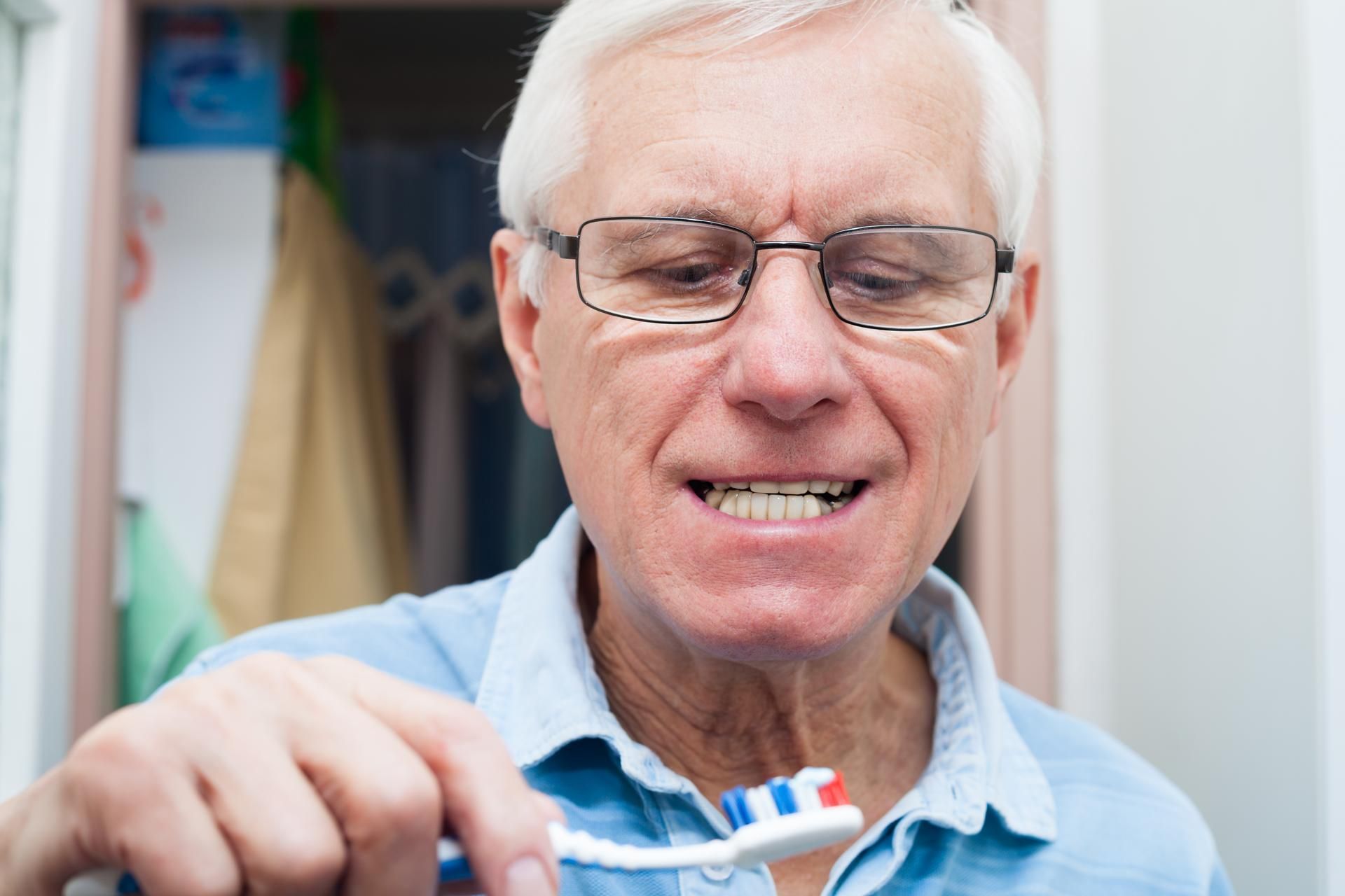 Man with glasses holding a toothbrush with toothpaste, grimacing in a bathroom.