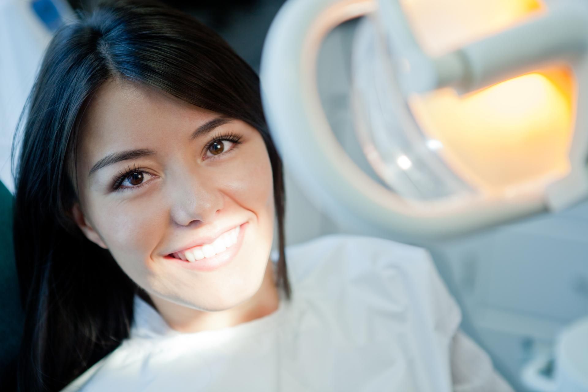 Woman in dentist's chair smiles, dental light overhead, white lab coat.