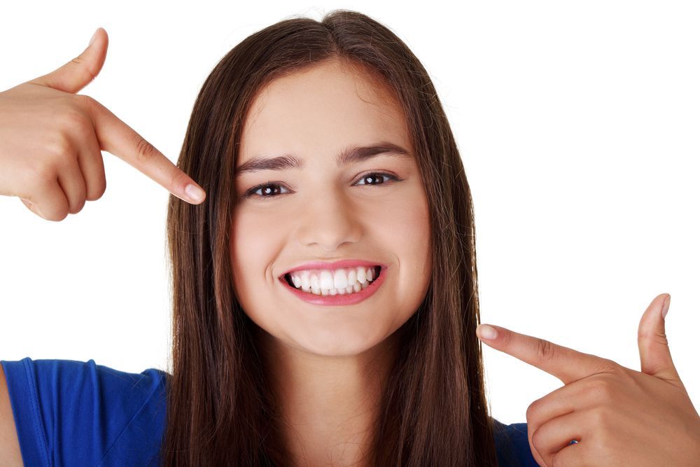 Woman smiling, pointing to her white teeth. Long brown hair, blue shirt, white background.