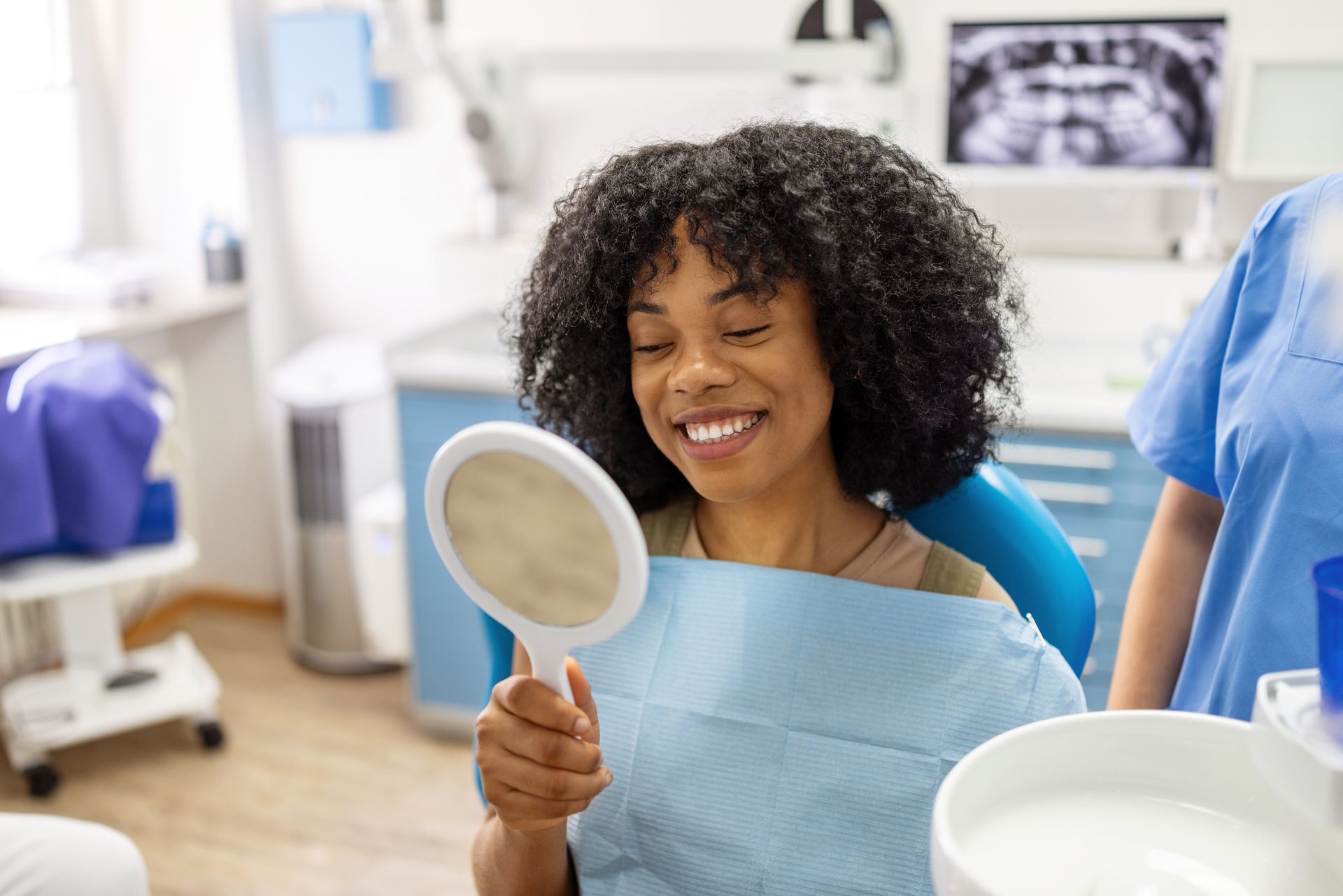 Woman smiling, looking at teeth in mirror after dental work in an exam room.