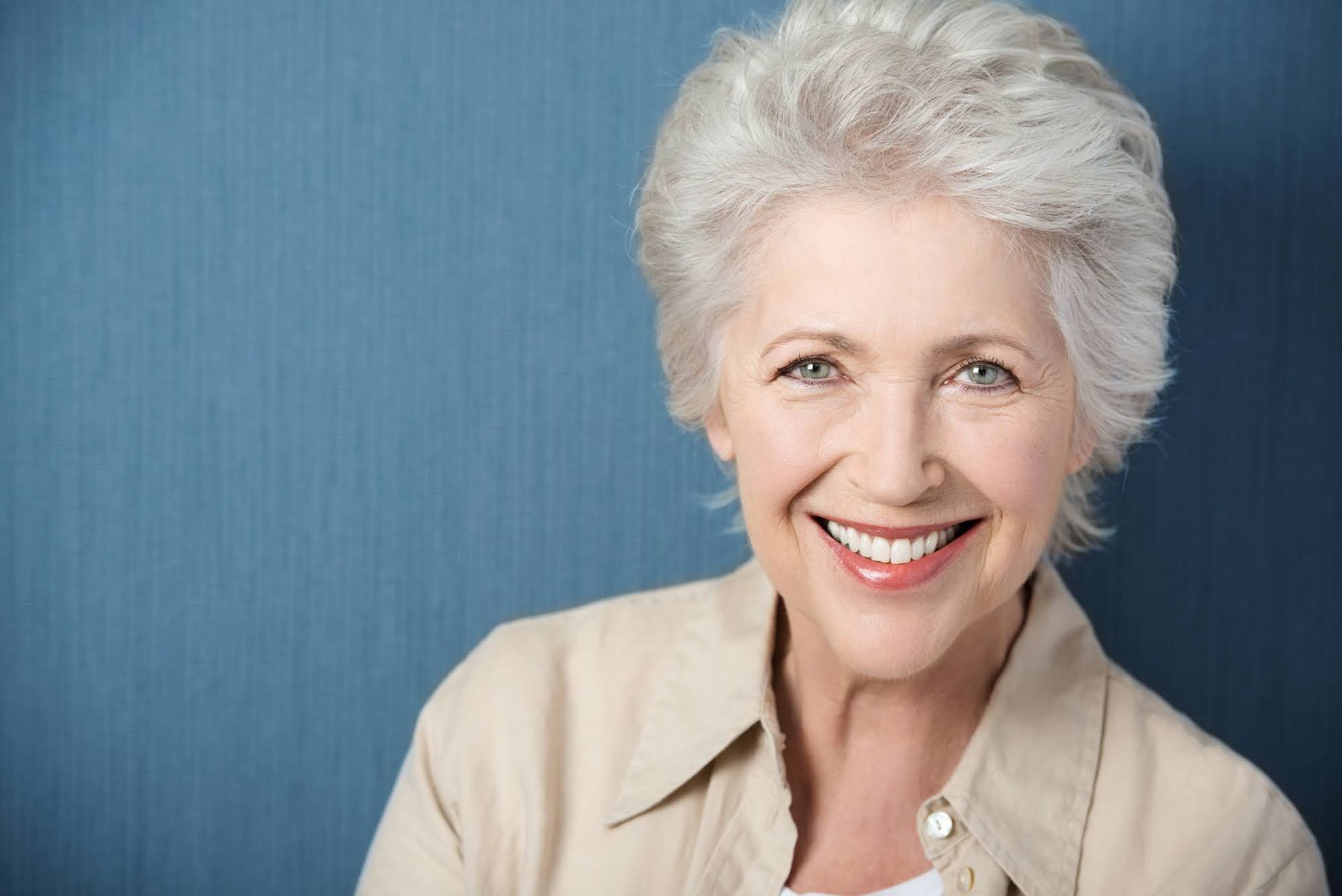 Woman with short, silver hair smiles at the camera, wearing a light-colored top, against a blue background.