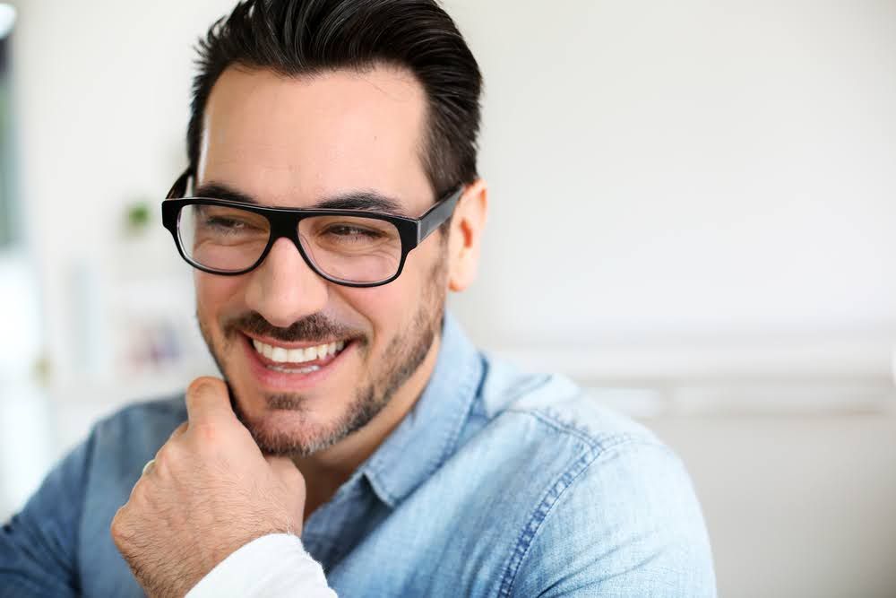 Man with glasses smiles, resting chin on hand. Wearing denim shirt, indoors.