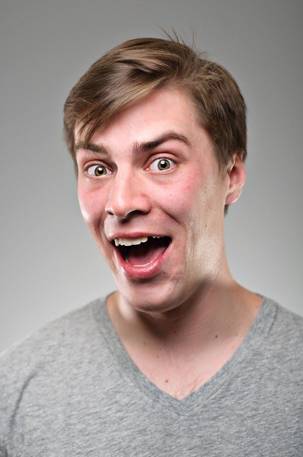 Man with wide eyes and open mouth, smiling. Wearing a gray v-neck shirt, against a gray background.