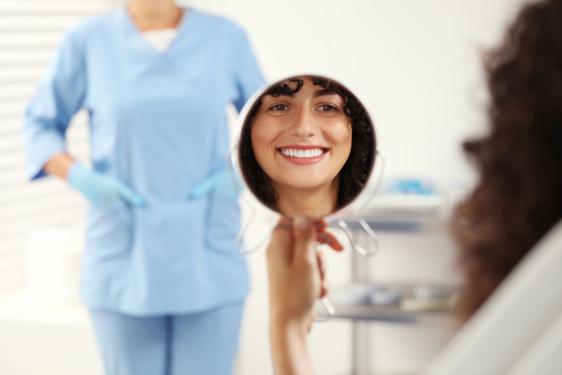 Smiling person looking at teeth in a mirror, dental assistant in blue scrubs visible.