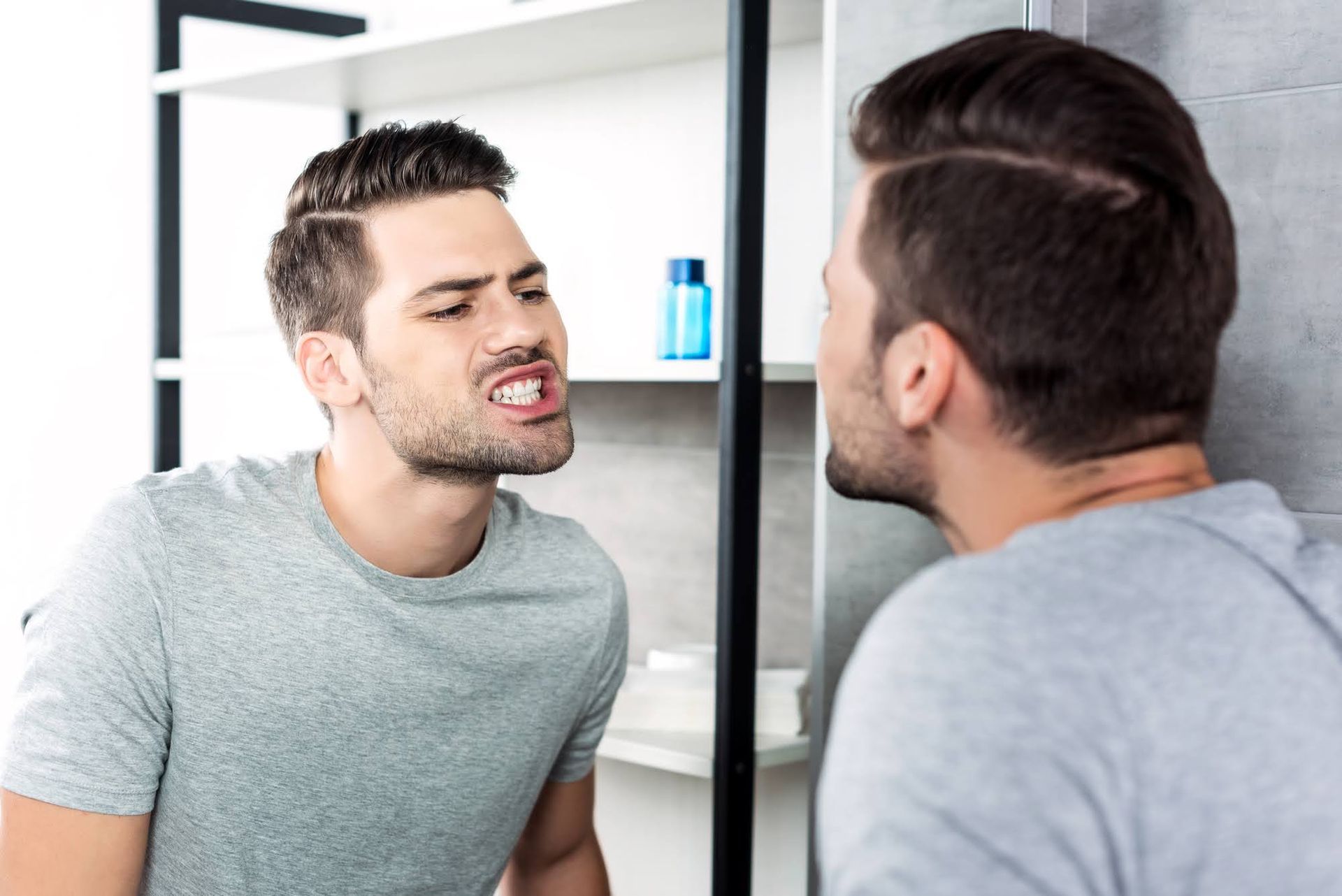 Man grimaces while looking at his reflection in a bathroom mirror.
