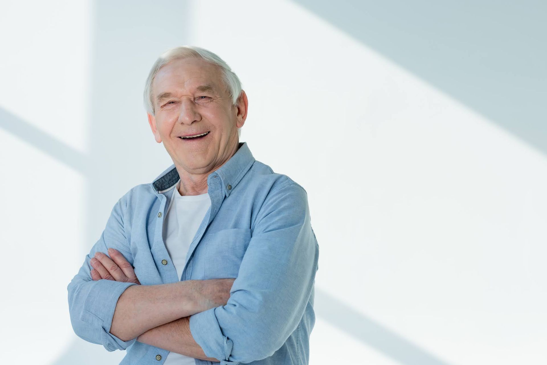Smiling older person with arms crossed, wearing a blue shirt and white t-shirt, standing in front of a white wall.