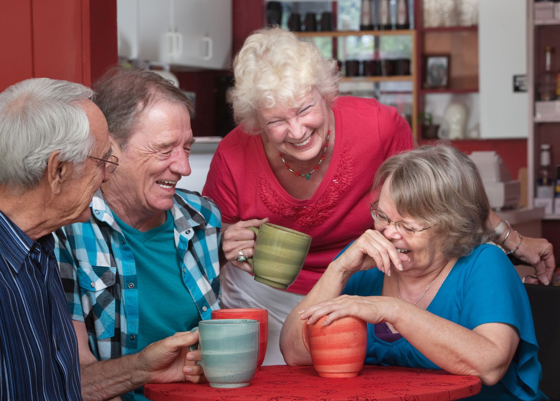 Four people laughing around a red table, holding mugs in a cafe setting.