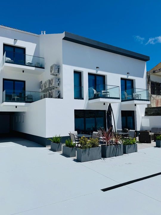 A modern white three-story building with blue balconies and large windows, featuring outdoor seating on a patio.