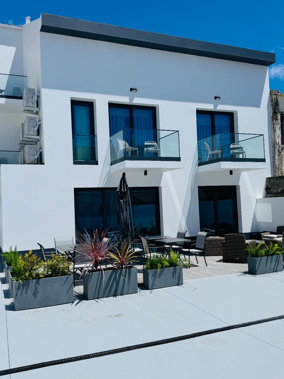 A modern white two-story building with two balconies, large windows, and outdoor seating on a patio with potted plants.