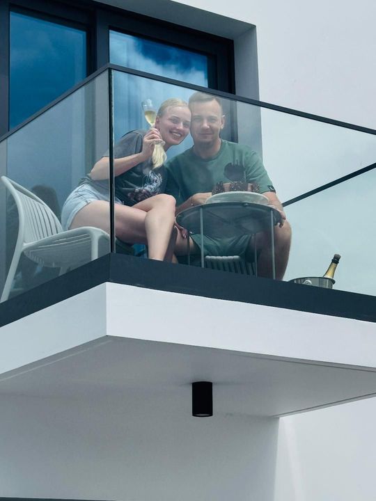 A couple sits on a modern balcony, smiling and holding drinks while sitting at a small table.