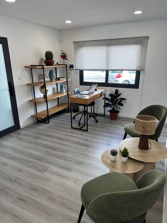 A small, minimalist office space with a desk, shelving unit, two green armchairs, and round coffee tables.