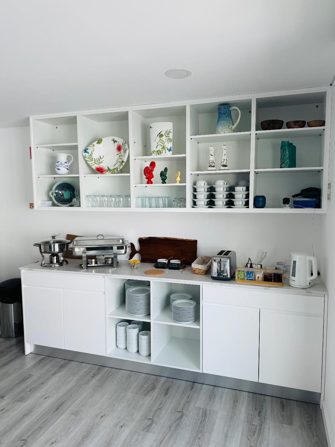 White kitchen cabinets and open shelving filled with dishware, glassware, and small appliances on a light wood floor.