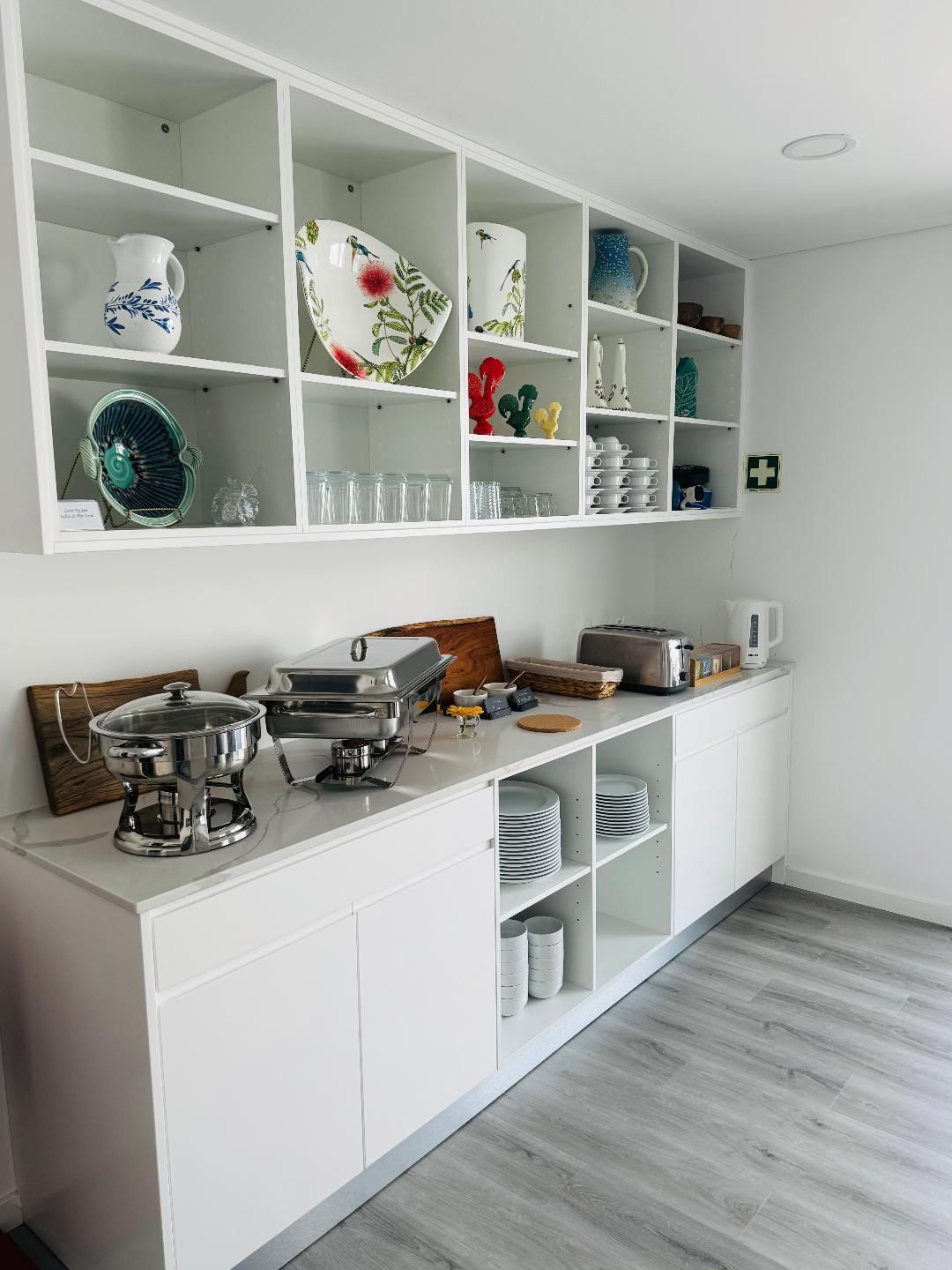 A white kitchen buffet with open shelving holding decorative plates, pitchers, and glasses, plus two metal food warmers.