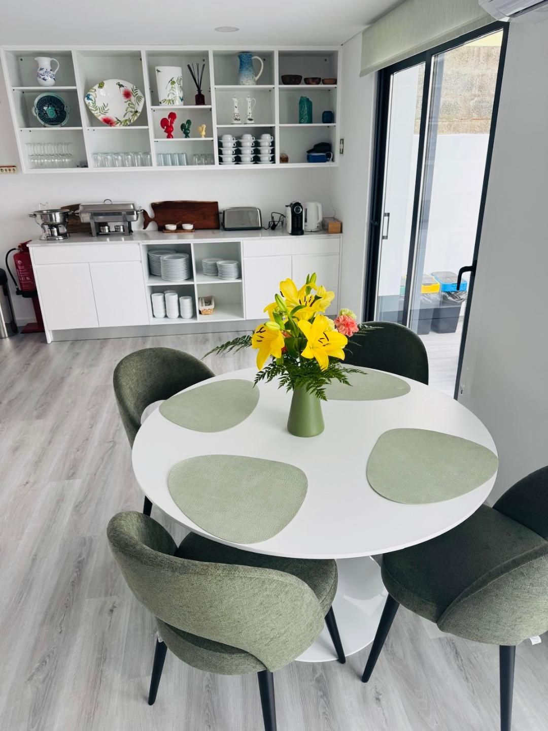 A dining area featuring a round white table with four sage green chairs, a vase of yellow lilies, and white wall shelving.