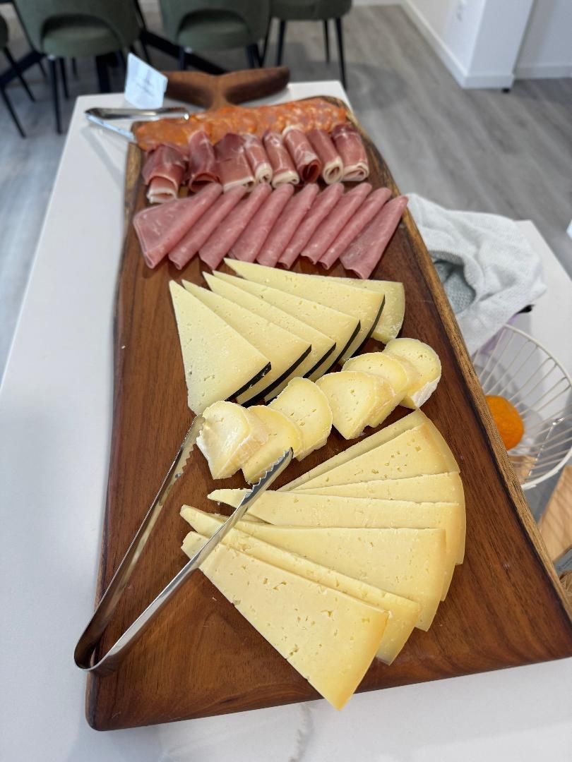 A wooden charcuterie board featuring rows of sliced cured meats and wedges of hard and soft cheeses, with metal tongs.