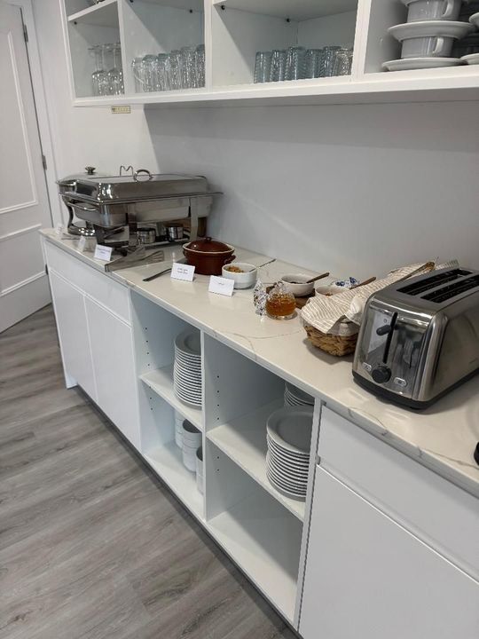 White kitchen counter with a buffet warmer, toaster, condiments, and dishware on shelves, in a bright, modern room.