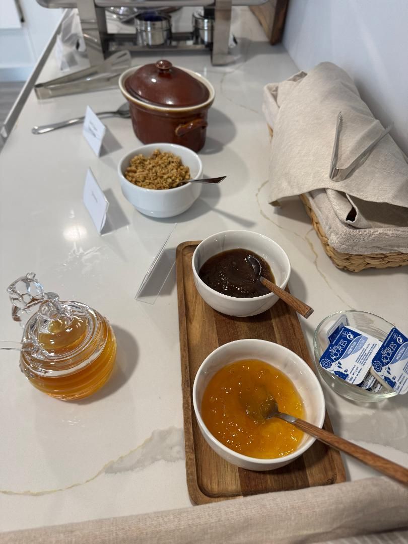 A breakfast buffet spread featuring honey, granola, jam, fruit preserves, and butter packets on a white countertop.