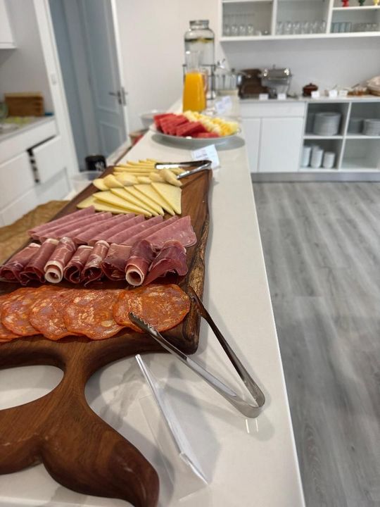 A long wooden serving board on a white counter holds rows of sliced meats and cheeses, with fruit in the background.