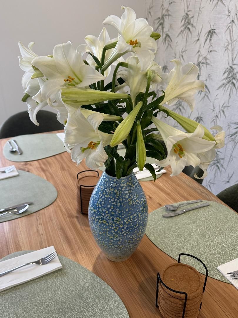 A bouquet of white lilies in a blue speckled vase, centered on a wooden dining table with place settings.
