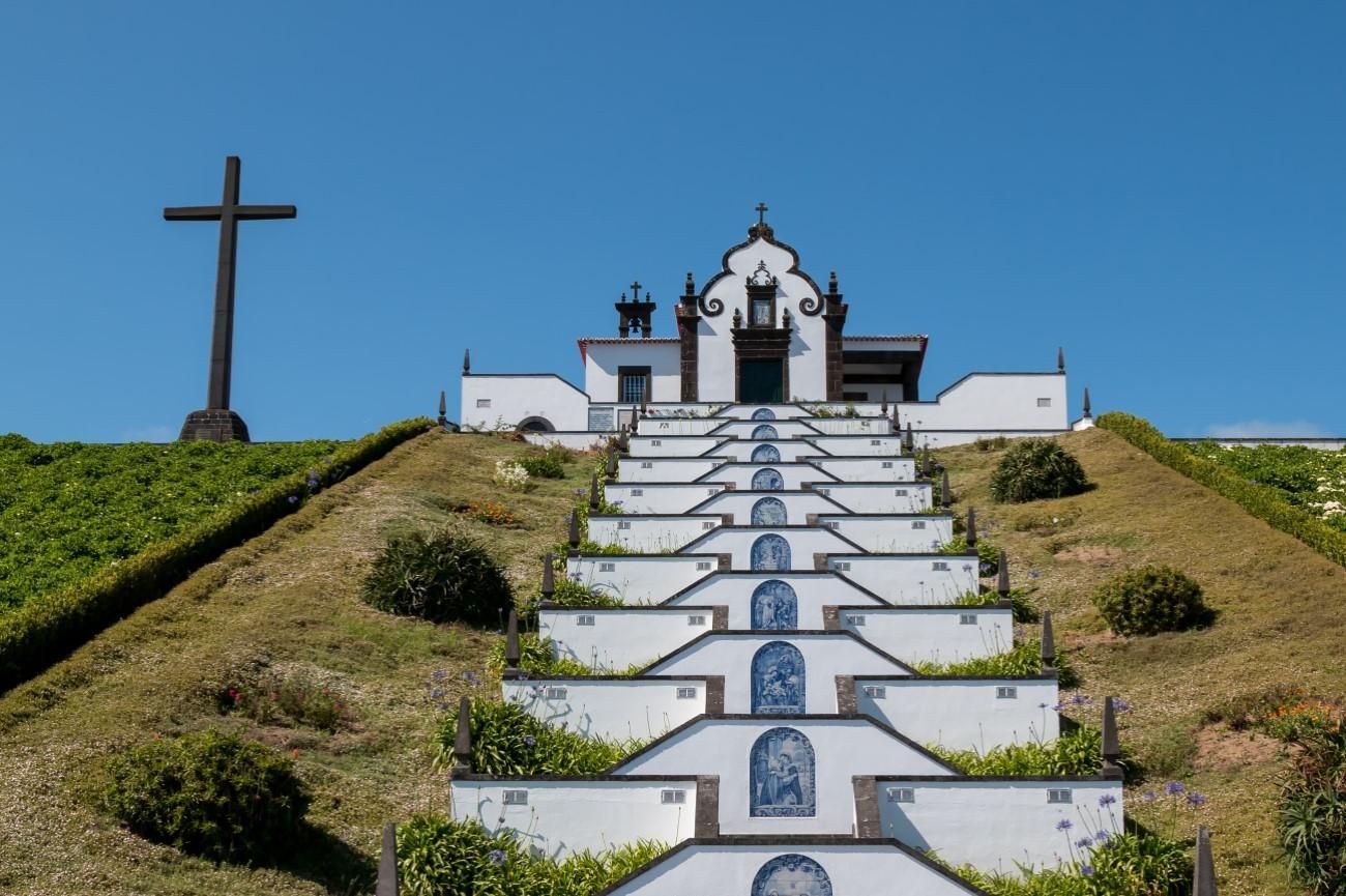 A white chapel sits atop a long, tiered stone staircase, with a tall dark cross on a grassy hill to the left.