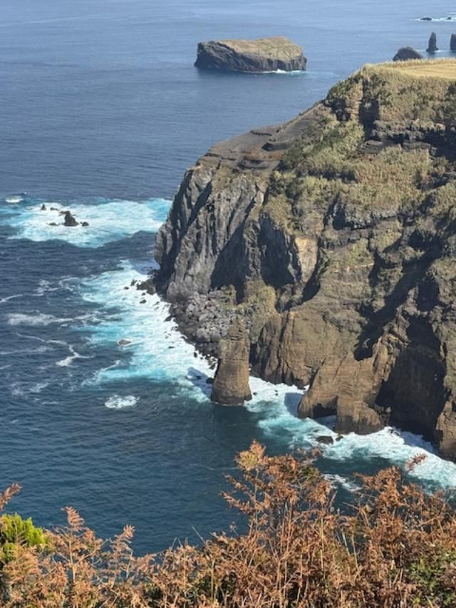 A rugged cliffside meets the ocean with white-capped waves, featuring a prominent sea stack and an island in the distance.