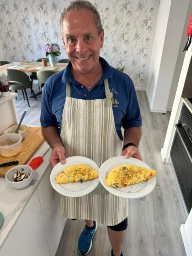 A person wearing an apron smiles while holding two plates, each containing a cooked omelet, in a kitchen setting.