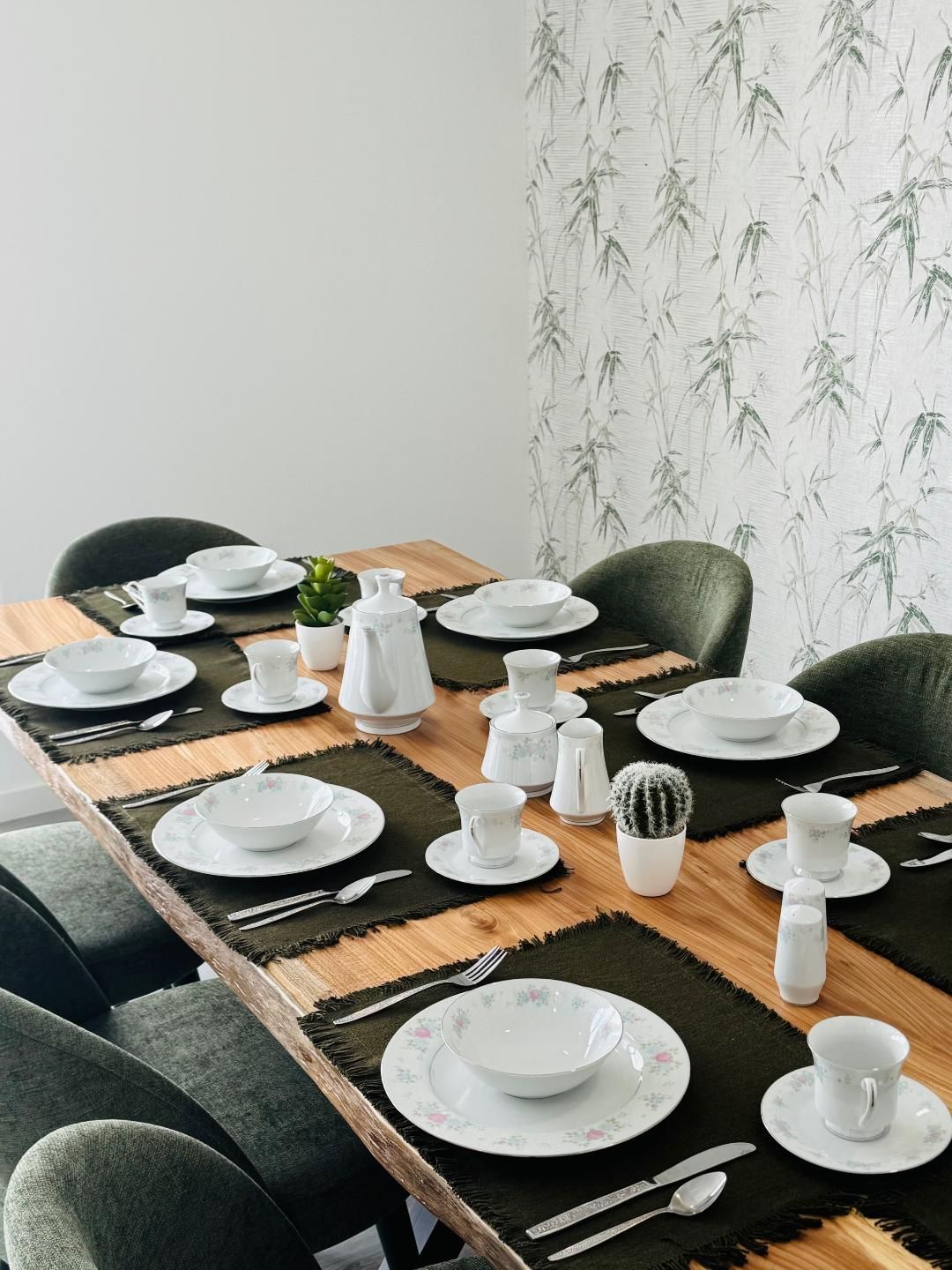 A wooden dining table set for six with white dinnerware, dark green placemats, and small potted plants against a wall.