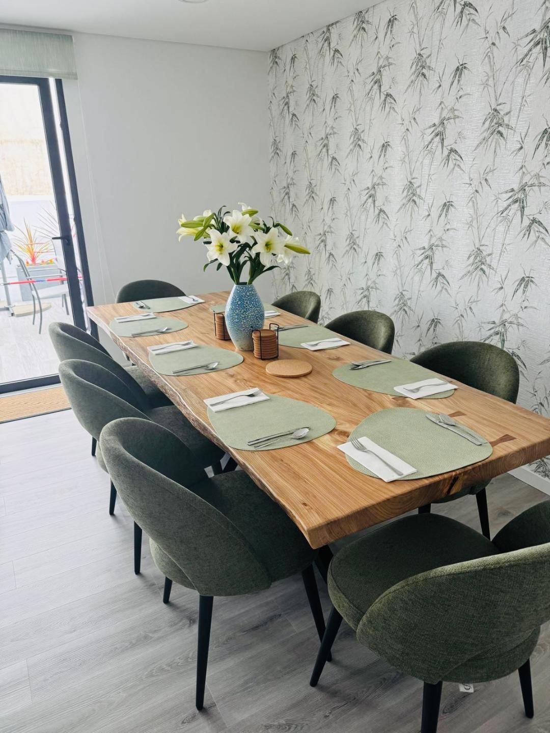 A wooden dining table with sage green chairs and floral wallpaper in a bright, modern dining room.
