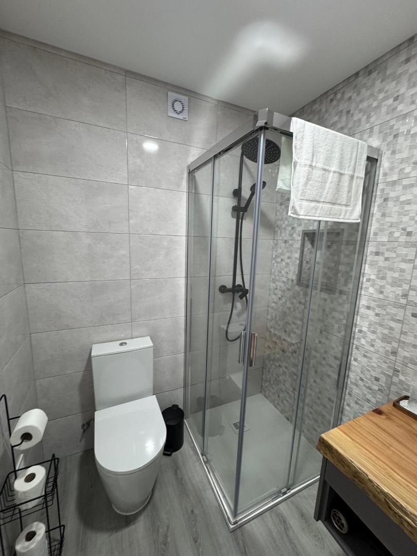 A modern bathroom with a white toilet, glass shower stall with black fixtures, and wood-topped vanity against tiled walls.