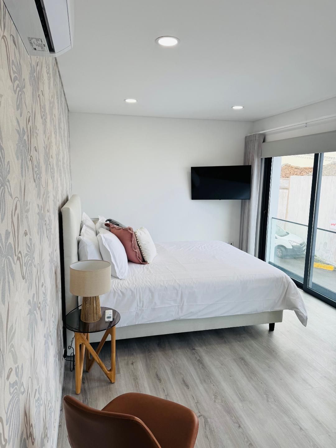 A bedroom featuring a bed with white bedding, patterned wallpaper, a bedside table with a lamp, and a wall-mounted TV.