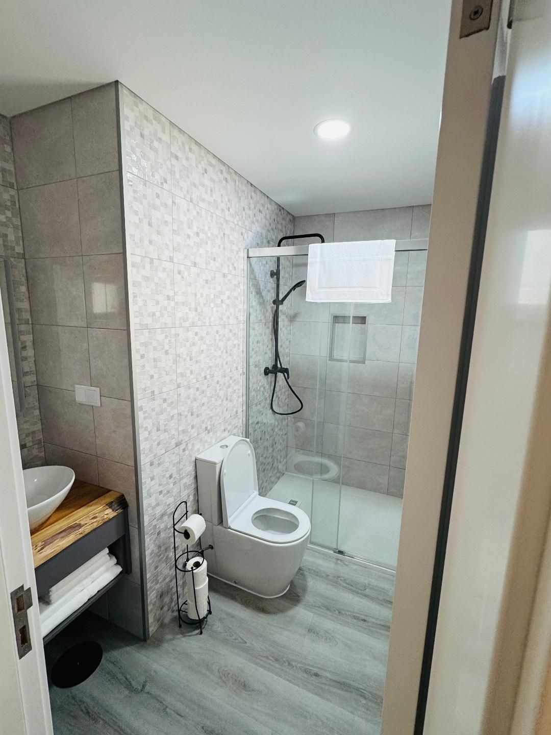 A modern bathroom with a wood-look vanity, white sink, toilet, and a glass-enclosed shower with tiled walls.