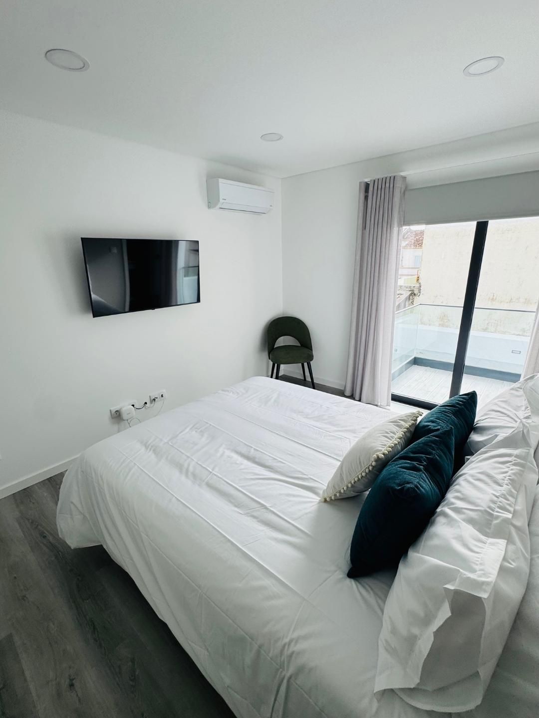 A modern, minimalist bedroom with a white bed, dark green pillows, wall-mounted television, and a sliding glass door.