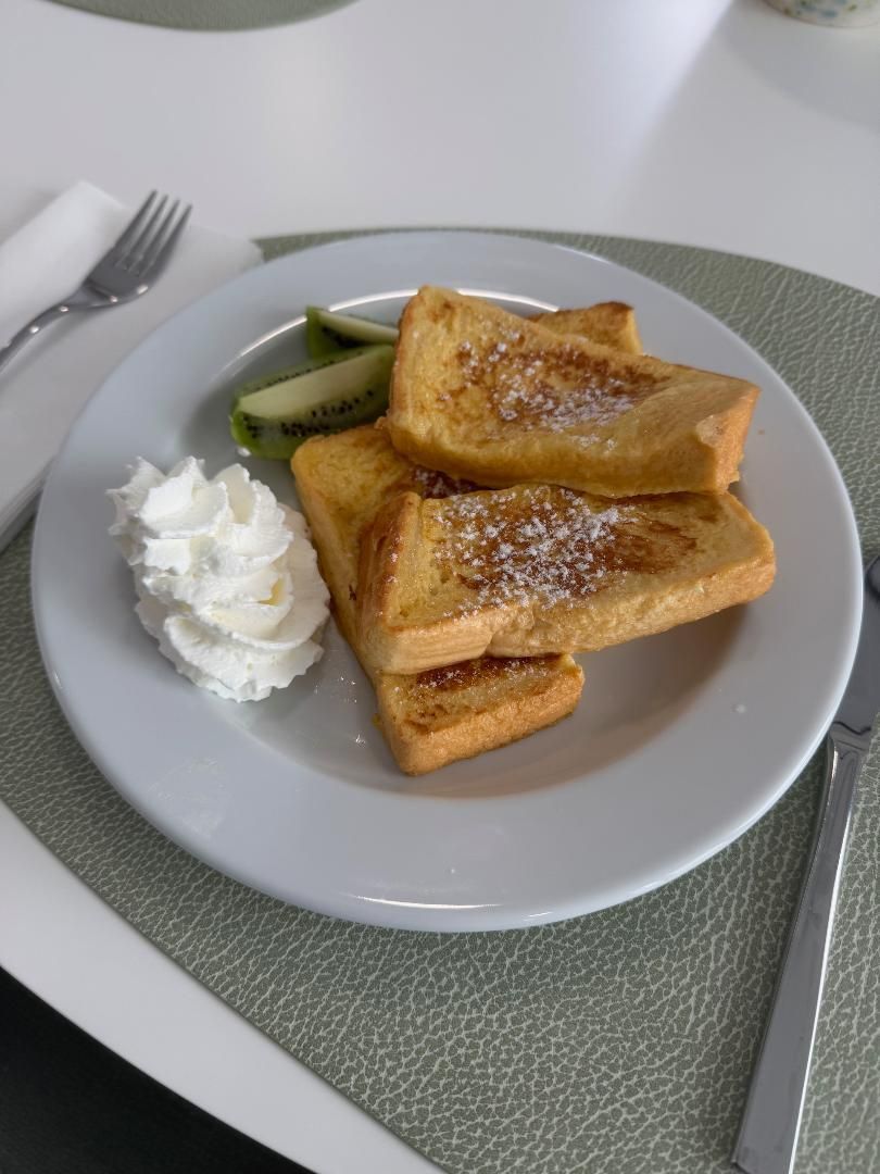 A plate of French toast sticks sprinkled with powdered sugar, served with a dollop of whipped cream and a slice of kiwi.