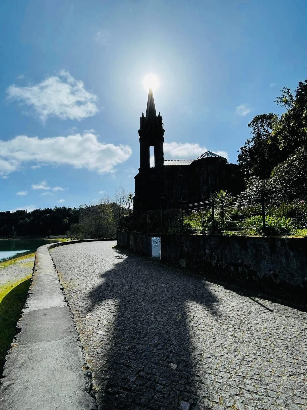Sunlight shines directly behind the tower of a dark stone church, casting a long silhouette across a gravel path.