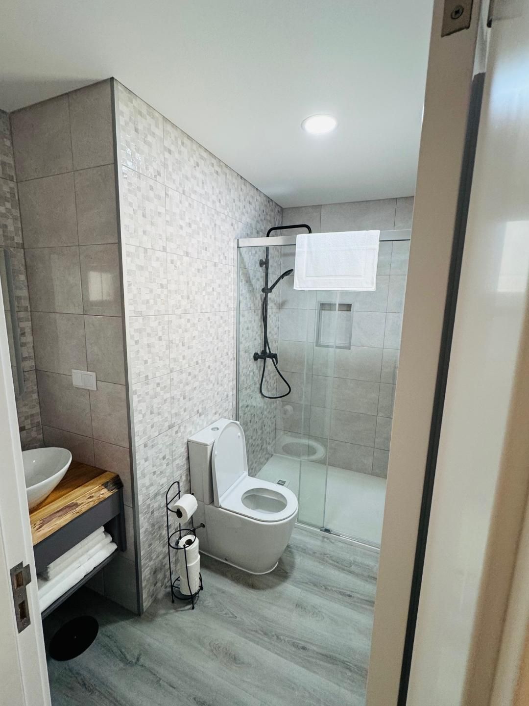 A modern bathroom with a wood-top vanity, a white toilet, and a glass-enclosed shower with tiled walls and gray flooring.