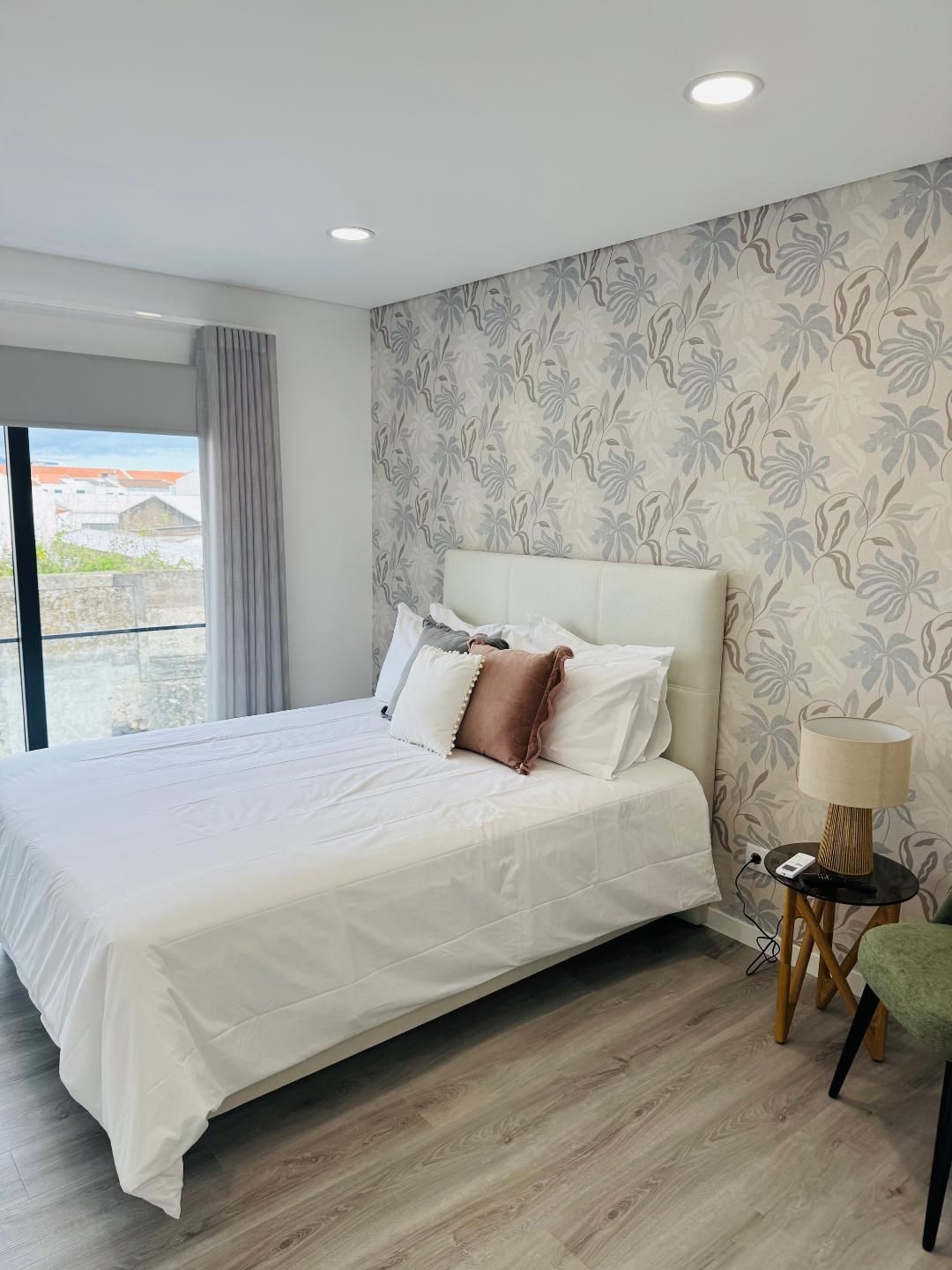 A modern bedroom with a white bed, patterned wallpaper, a side table with a lamp, and a window with grey curtains.