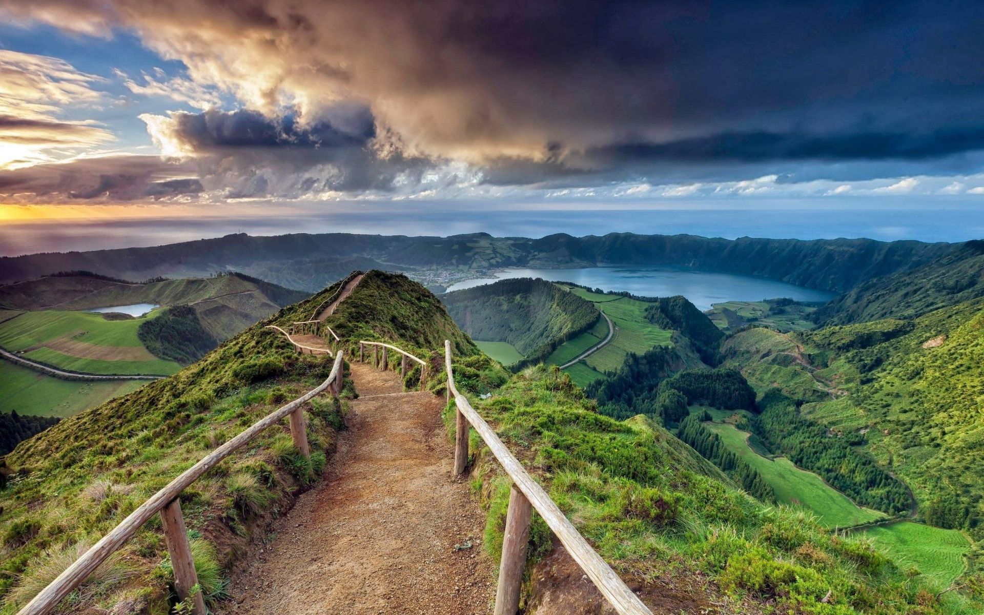 A dirt hiking path with a wooden railing leads toward a vast, green volcanic crater lake under a dramatic, cloudy sky.