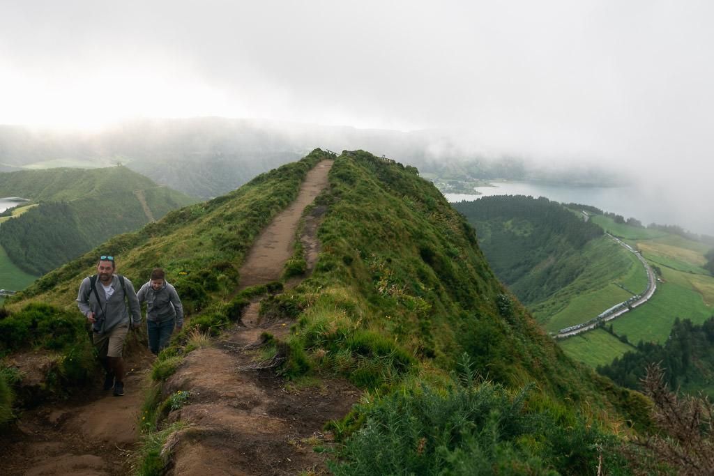 Two people hike along a narrow, grassy ridge trail overlooking a lush valley and lakes under a cloudy sky.