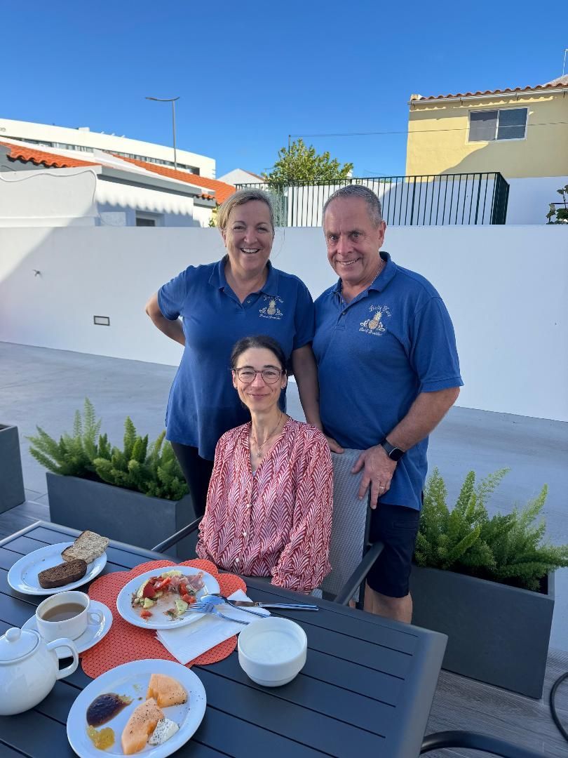 Three people pose outdoors on a sunny terrace at a breakfast table with plates of food and drinks.