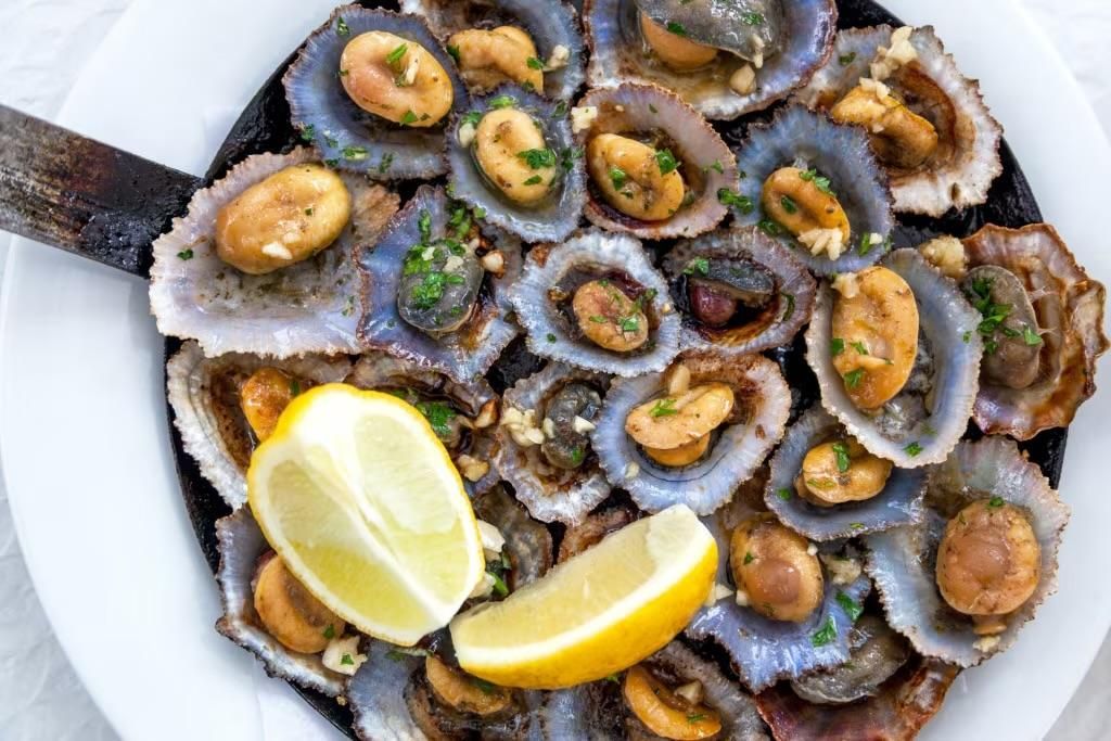 A top-down view of grilled limpets served in a dark iron pan with lemon wedges on a white plate.