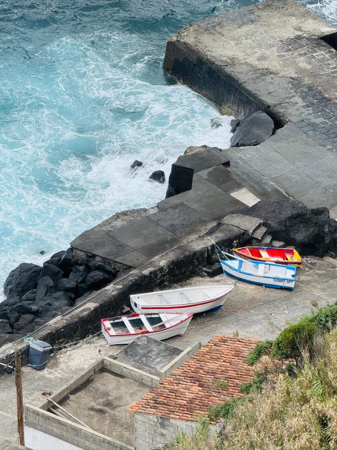 Three small boats in white, red, and blue rest on a concrete landing beside churning turquoise ocean waves.